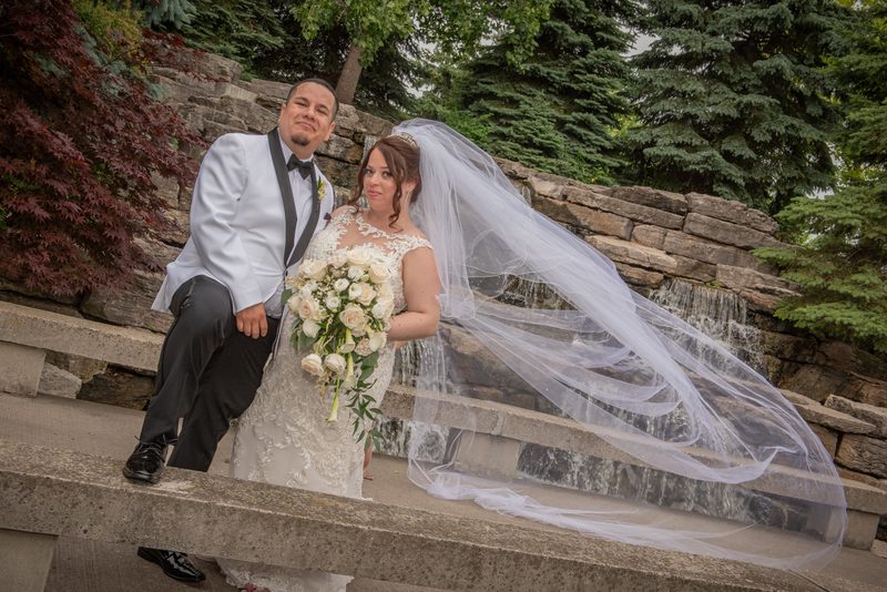 A bride and groom pose for a portrait on stone steps beside a small waterfall, surrounded by lush greenery.