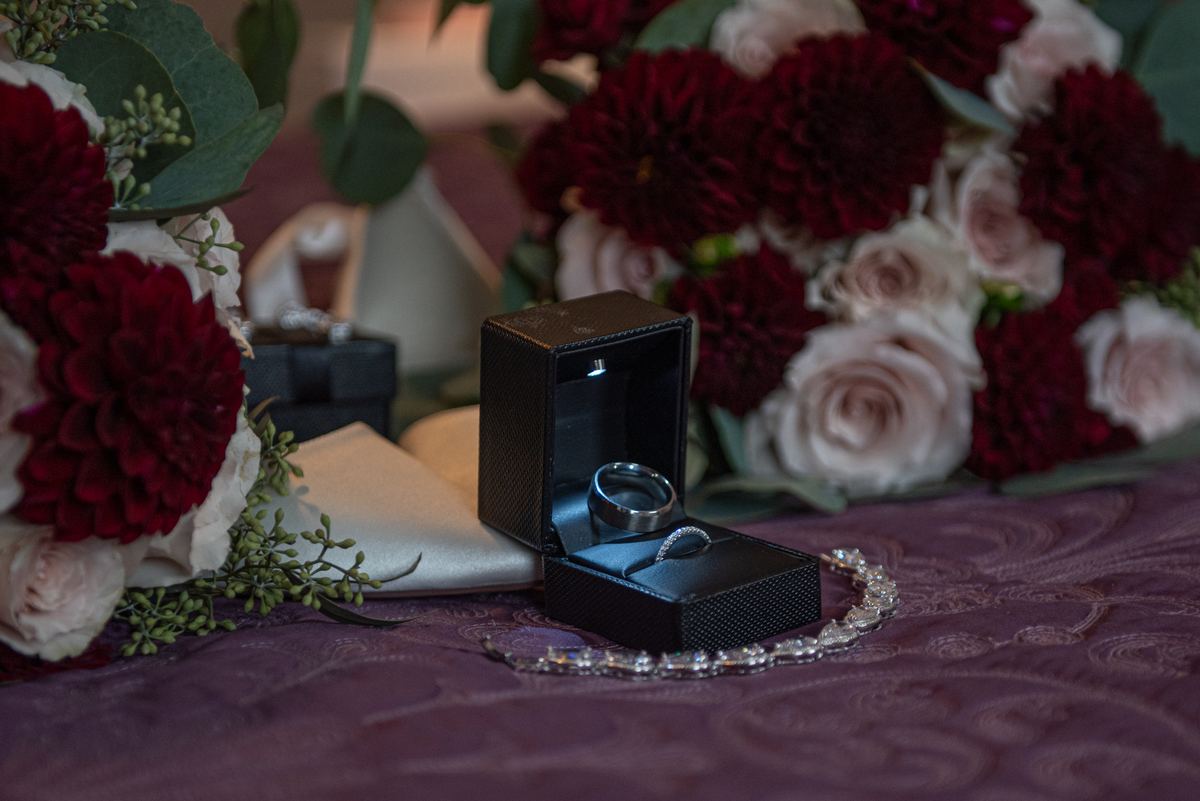 Wedding ring in an open black velvet box with a sparkling bracelet and floral arrangements, bathed in soft light.