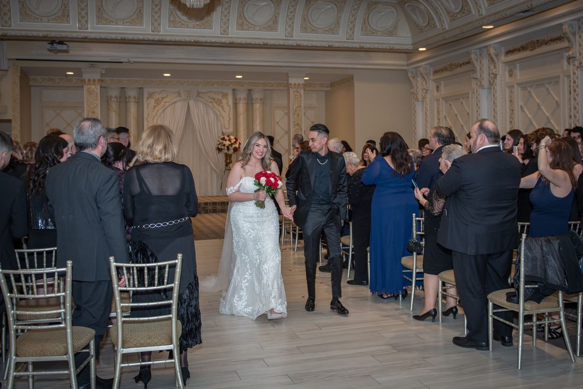 A bride and groom walk down the center of an ornate ballroom, smiling at guests as they proceed toward the ceremony area.