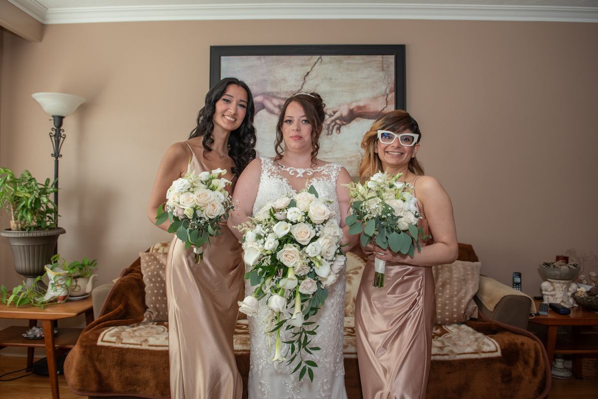A bride and two bridesmaids pose together indoors, their elegant gowns and floral bouquets highlighted by soft light.