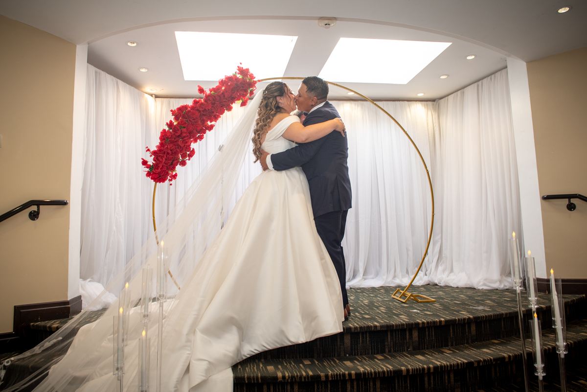 Bride and groom kiss on an indoor staircase under a golden arch covered in red flowers, surrounded by white drapes.