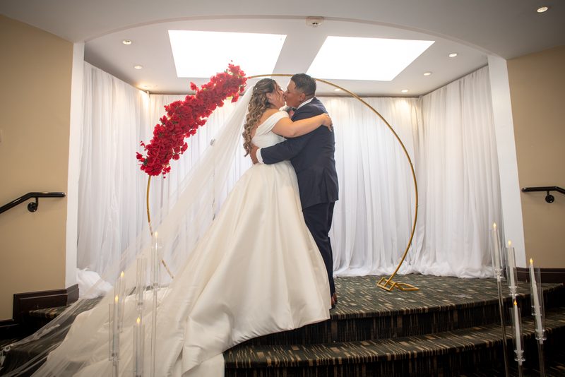 Bride and groom kiss on an indoor staircase under a golden arch covered in red flowers, surrounded by white drapes.