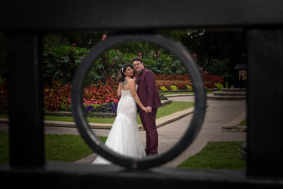 Newlyweds embrace through a circular fence opening in a garden, illuminated by soft light and surrounded by autumn foliage.