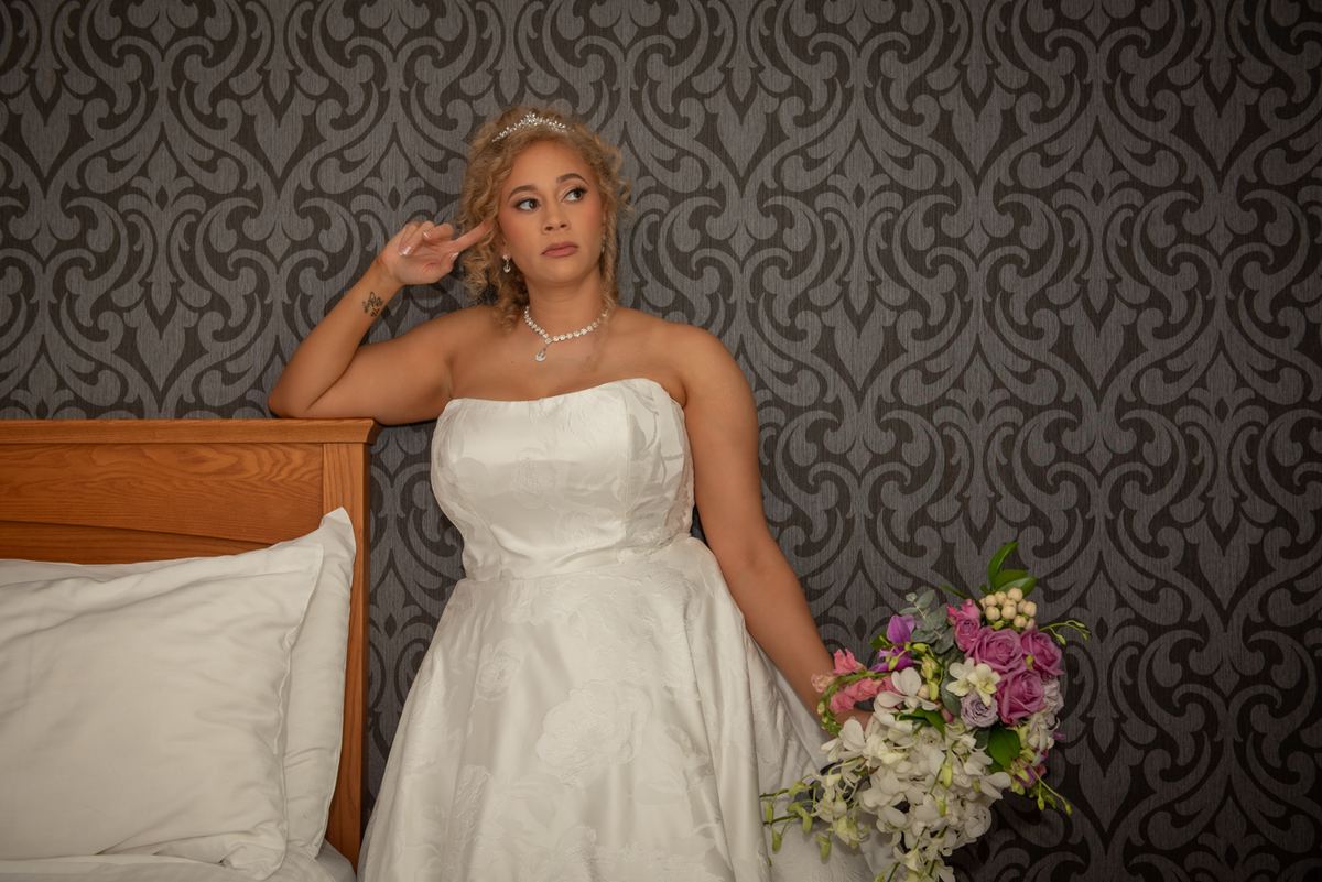 A bride in a white gown leans against patterned wallpaper, holding flowers and gesturing playfully with her finger.