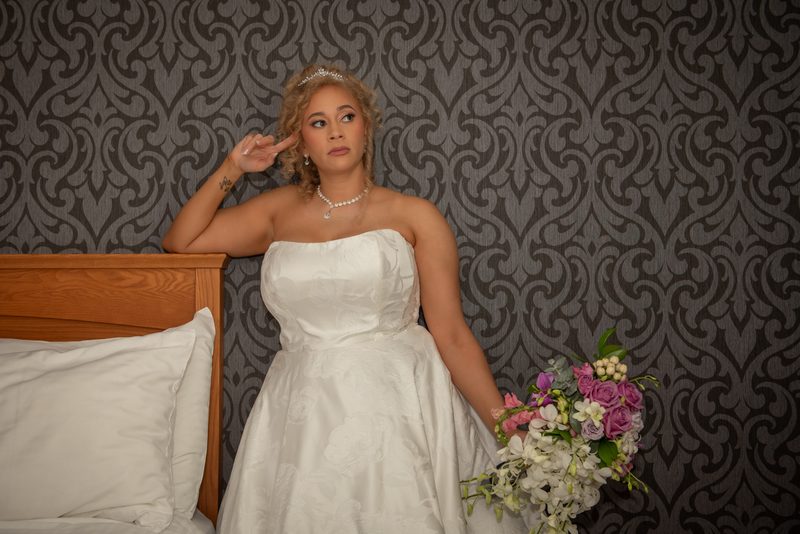 A bride in a white gown leans against patterned wallpaper, holding flowers and gesturing playfully with her finger.