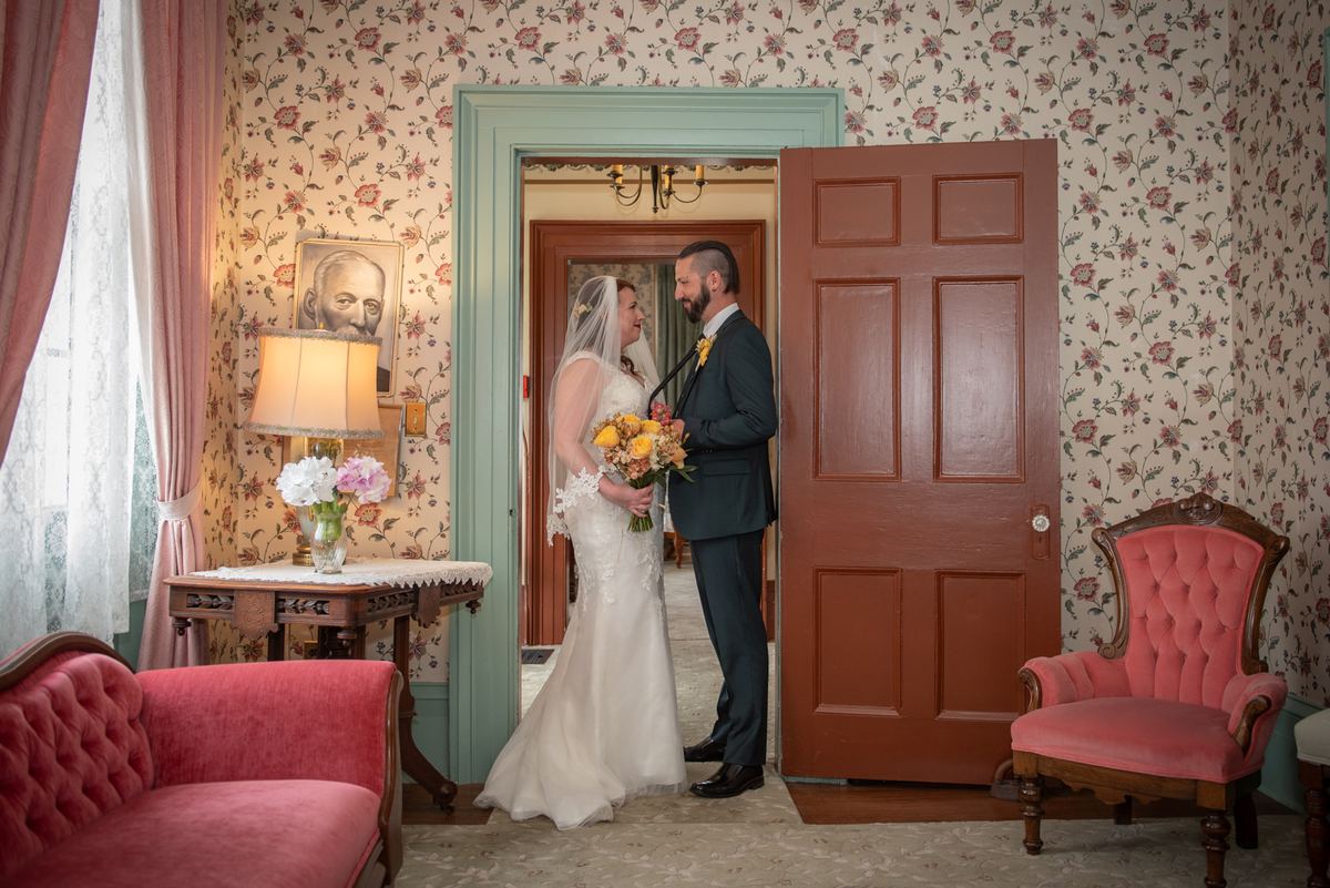 Bride in white gown and veil holding flowers meets groom in dark suit for first look in warmly lit vintage room.