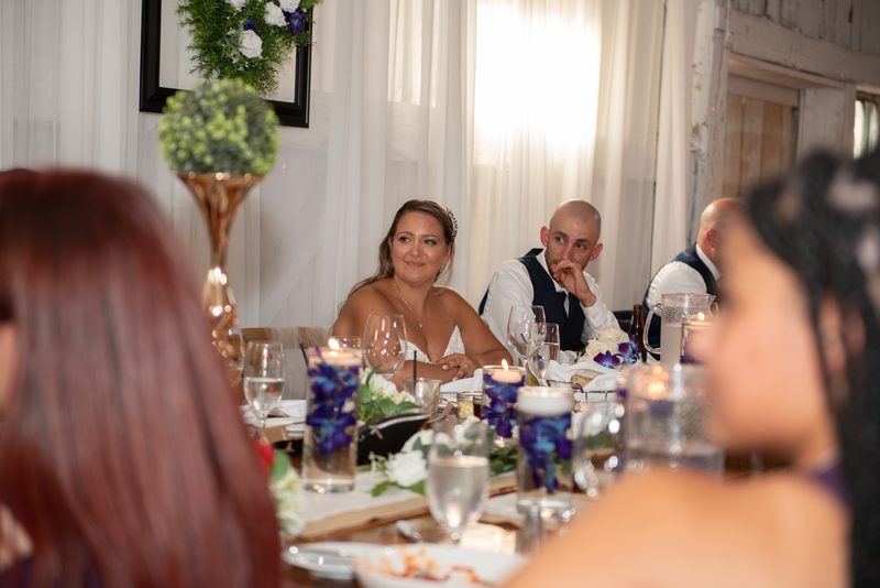 Bride and groom sit at a decorated table during a wedding reception, smiling as guests converse around them.