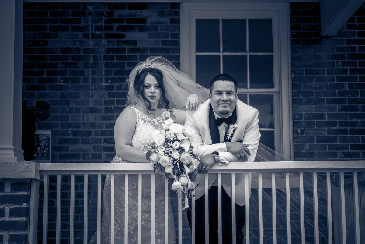 A newlywed couple in formal attire pose on a porch railing, the bride with a bouquet, under cool blue monochrome lighting.