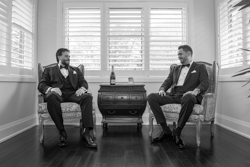 Two men in tuxedos sit and converse warmly, illuminated by soft light from shuttered windows, with a wine bottle nearby.