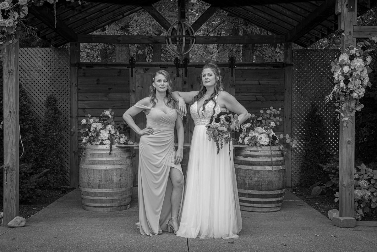 Two women in wedding and formal gowns pose confidently under a rustic wooden arbor adorned with flowers.