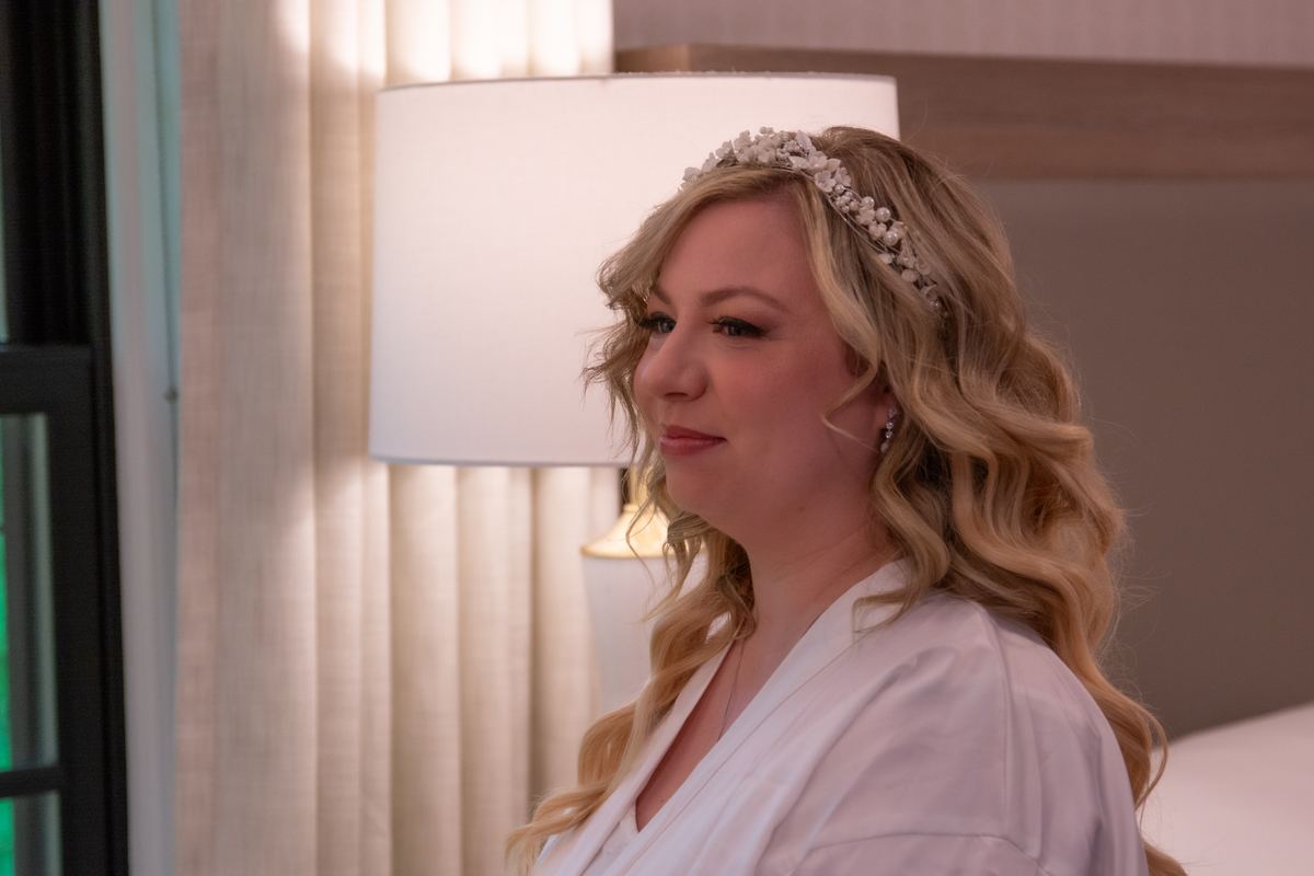 A bride in a white robe and floral headpiece stands by a lamp, her thoughtful gaze away from the camera.