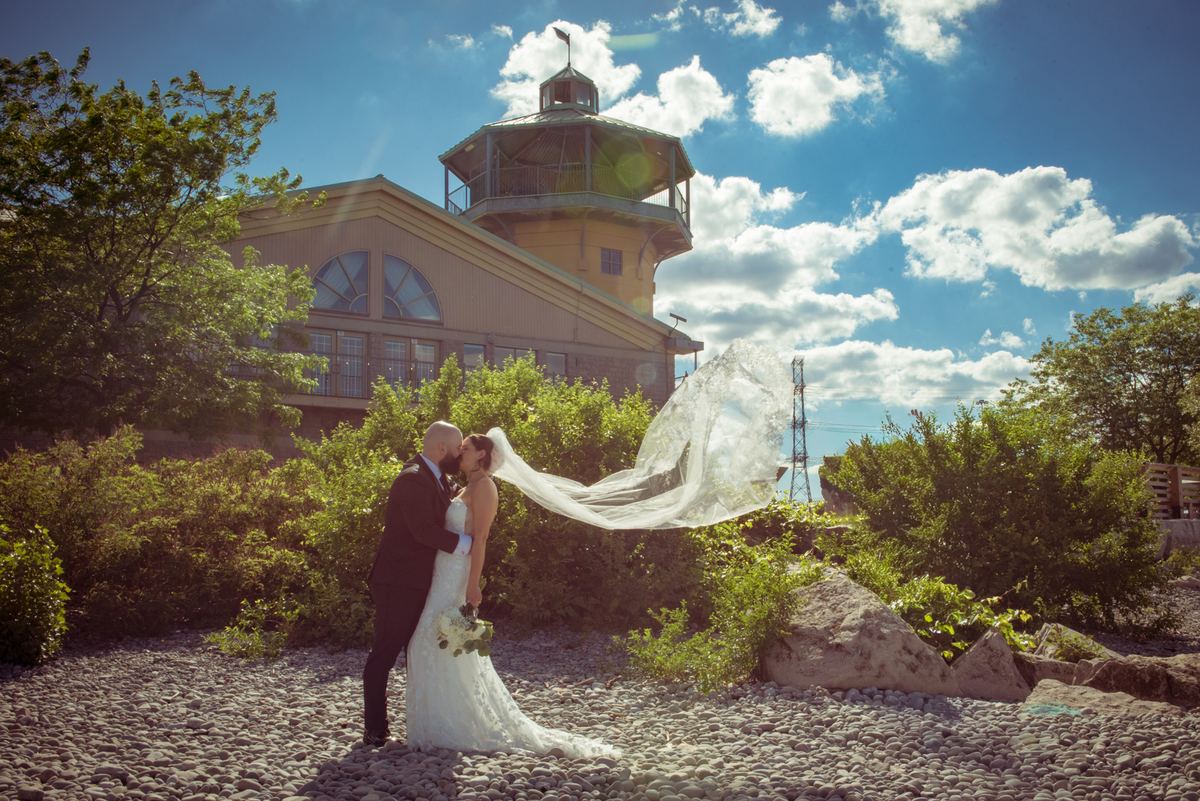 Bride and groom kiss on a pebbled shore, surrounded by greenery and an octagonal tower under a sunny sky.