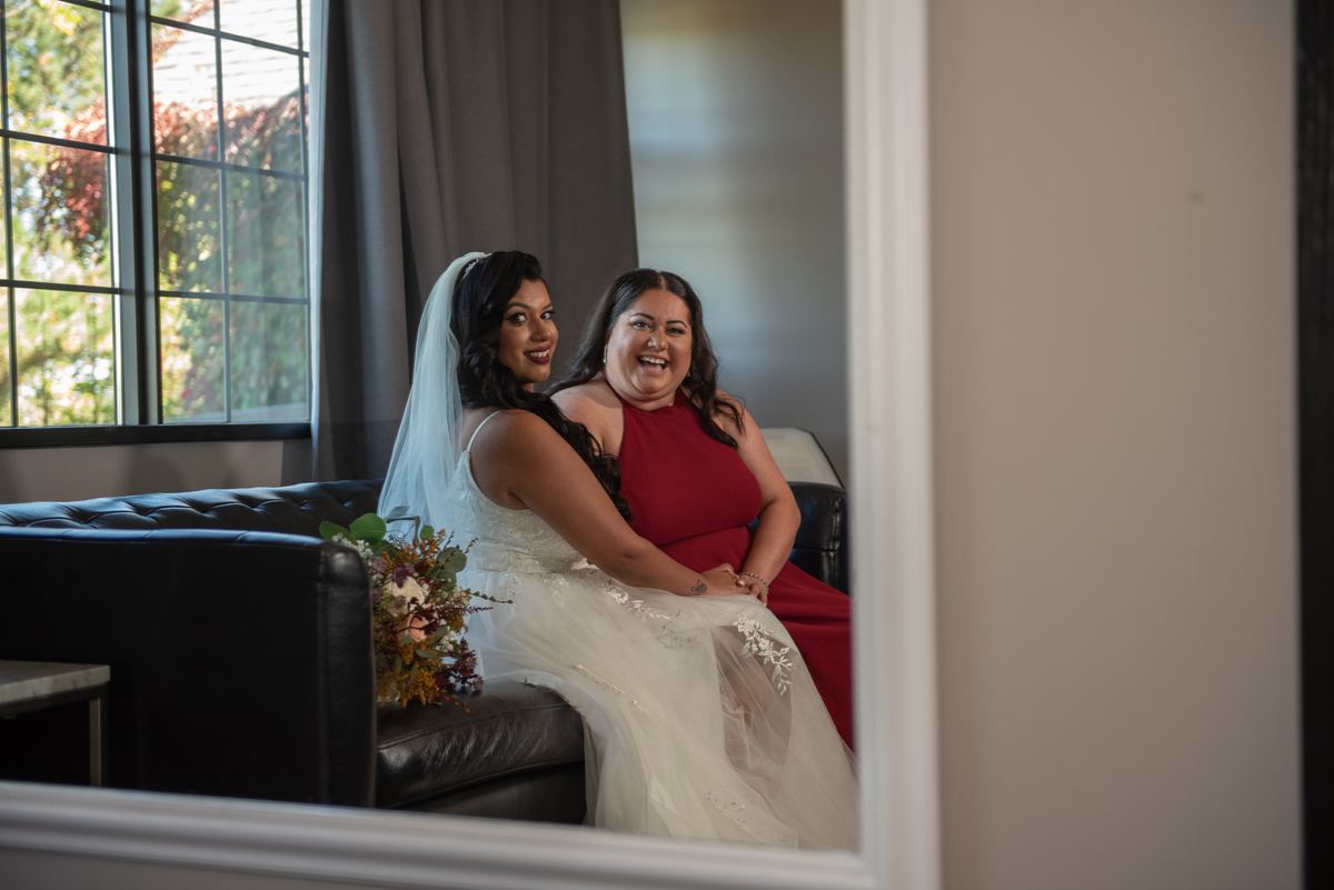 Two women, one in a wedding gown and the other in a red dress, sit on a black leather couch with bright smiles.