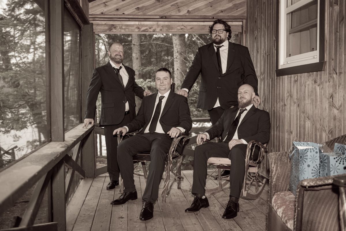 Four formally dressed men pose together on a rustic wooden porch surrounded by trees, one standing behind the others.