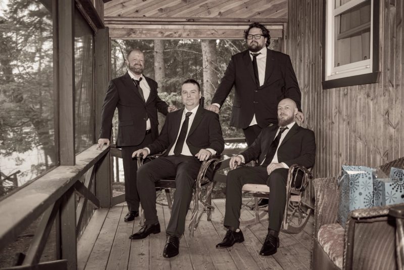 Four formally dressed men pose together on a rustic wooden porch surrounded by trees, one standing behind the others.
