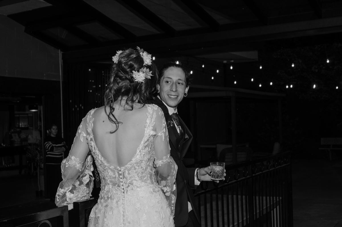 A bride in an ornate lace gown and floral hairpieces stands with her smiling groom, who holds a drink.