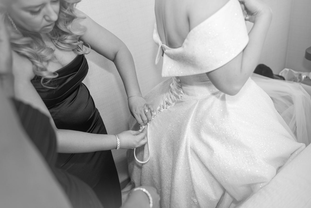 A bridesmaid fastens the lacing on a bride's sparkling wedding gown in an intimate moment of preparation.