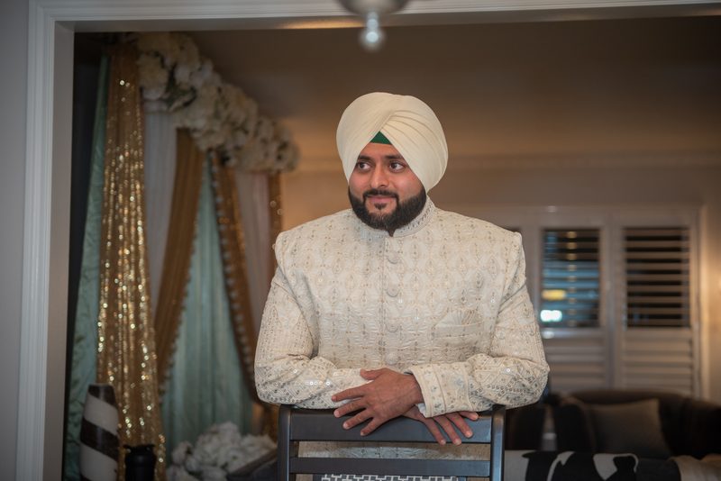 A groom in a cream sherwani and white turban with a green band stands thoughtfully near sequined drapes.