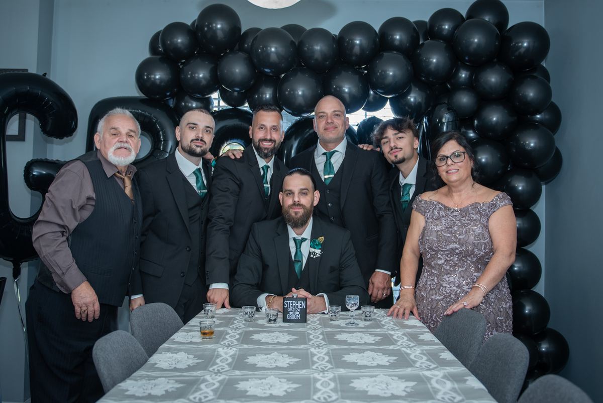 A groom and his family and friends pose together in front of a black balloon arch at a decorated table.