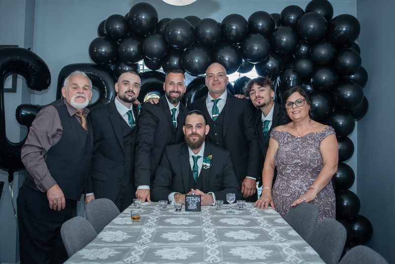 A groom and his family and friends pose together in front of a black balloon arch at a decorated table.