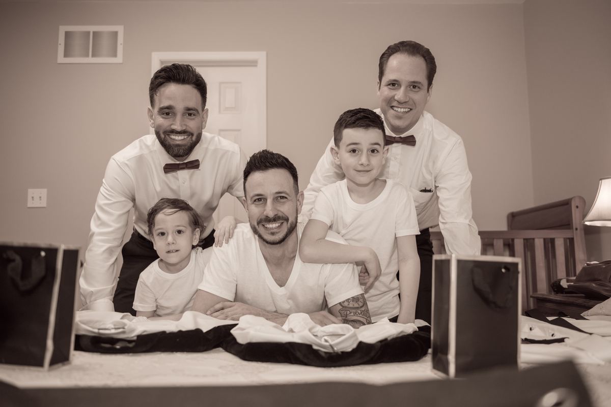 Groom and groomsmen with two boys around a bed of folded shirts and gift bags during wedding preparations.