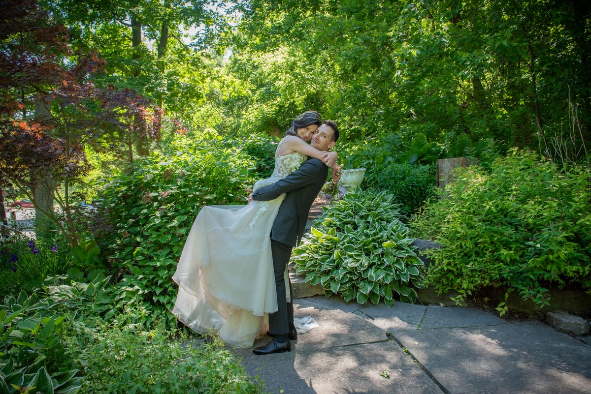 A groom lifts his bride in a celebratory dip in a sun-dappled garden, their faces close with affection.
