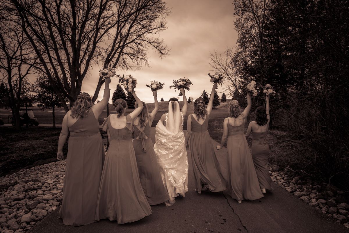 A bride and her bridesmaids walk along a path, raising bouquets in celebration under the soft light of early evening.