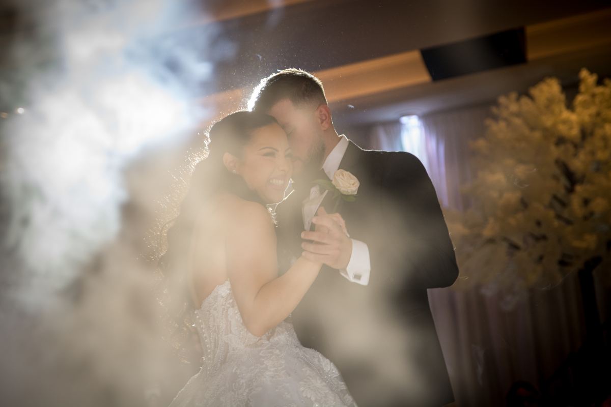 Bride and groom embrace joyfully during first dance, faces glowing with backlight, particles floating in the light.