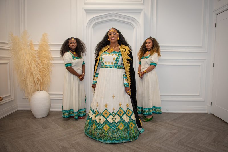 Three women in traditional attire stand in an elegantly paneled room; the central figure smiles warmly in her ornate gown.