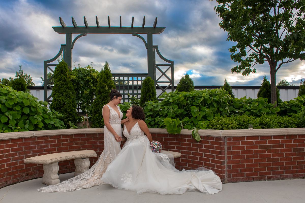 Two brides smile and touch gently under a pergola surrounded by greenery during a wedding photoshoot.