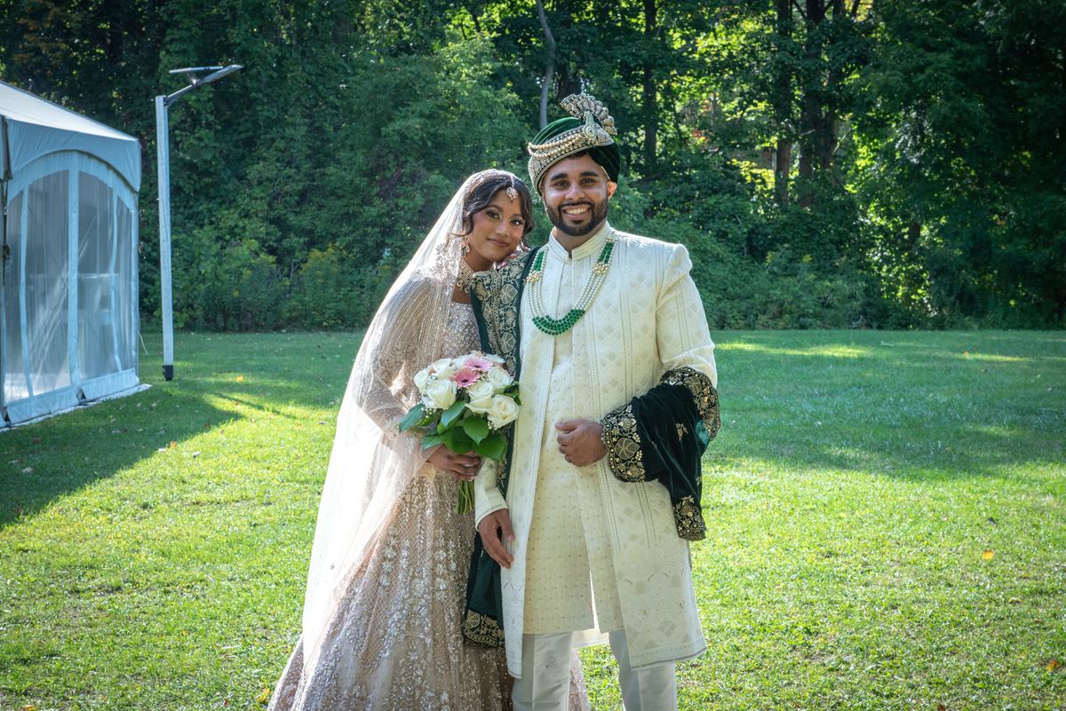 A smiling South Asian bride in a shimmering lehenga and groom in an ornate sherwani stand together on a sunlit lawn.