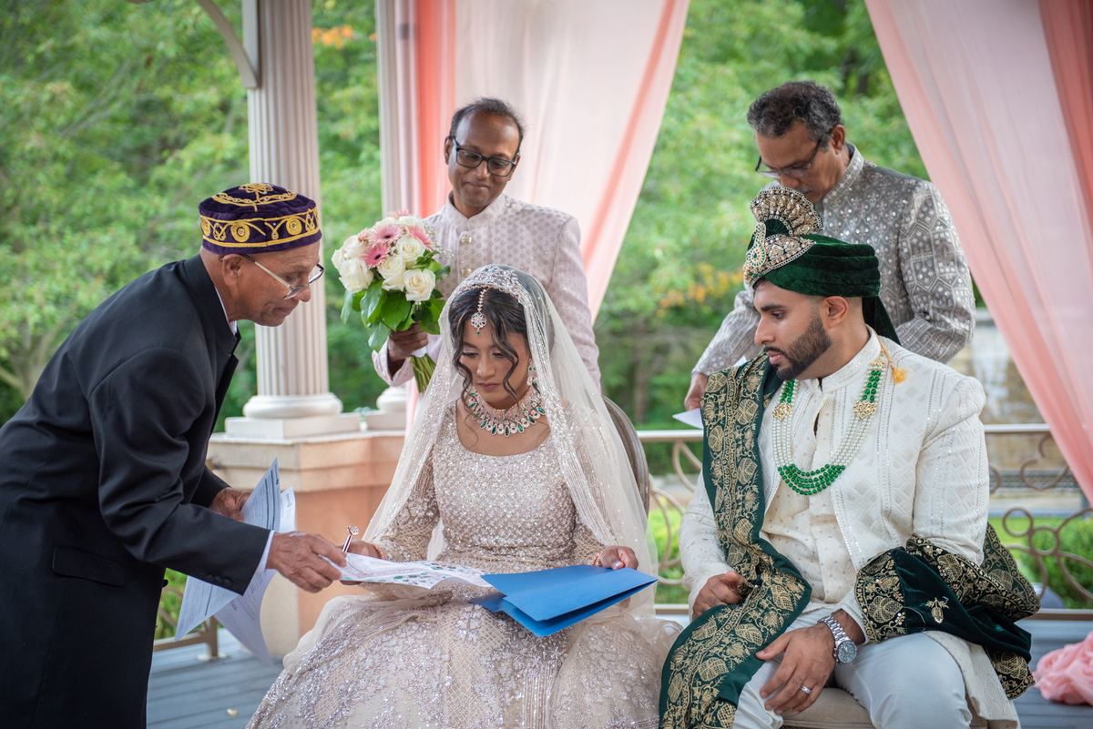 A bride in white and groom in traditional attire sign documents under a canopy during an outdoor ceremony.