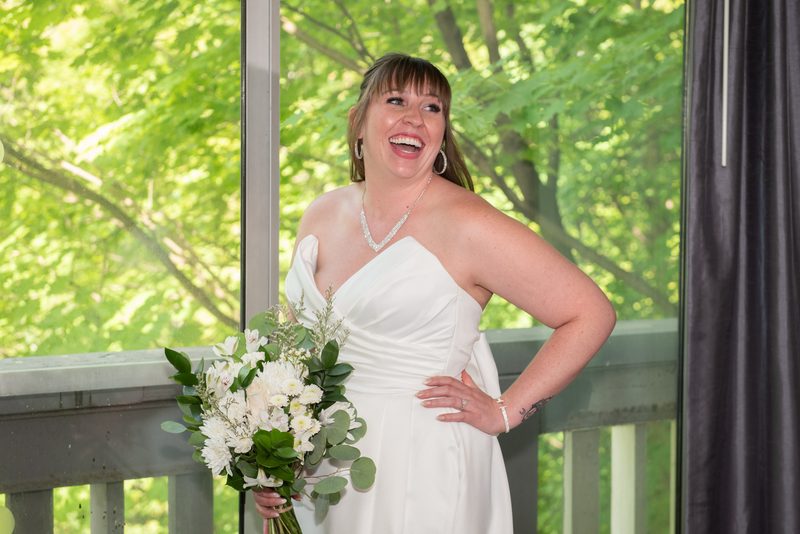A bride in a white strapless gown laughs by a window with lush green trees outside, holding a bouquet of white flowers.