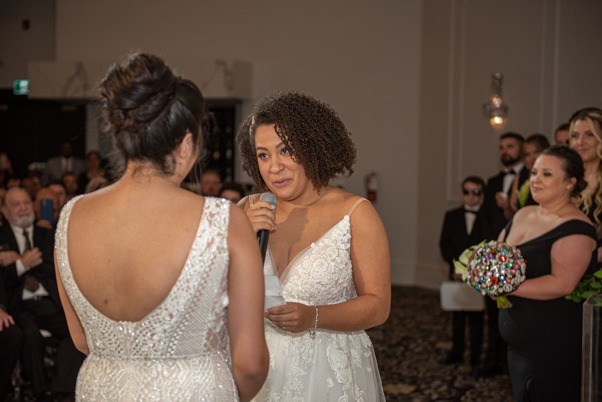 A bride in a sparkling gown faces her partner in a lace dress holding a microphone during a wedding toast.