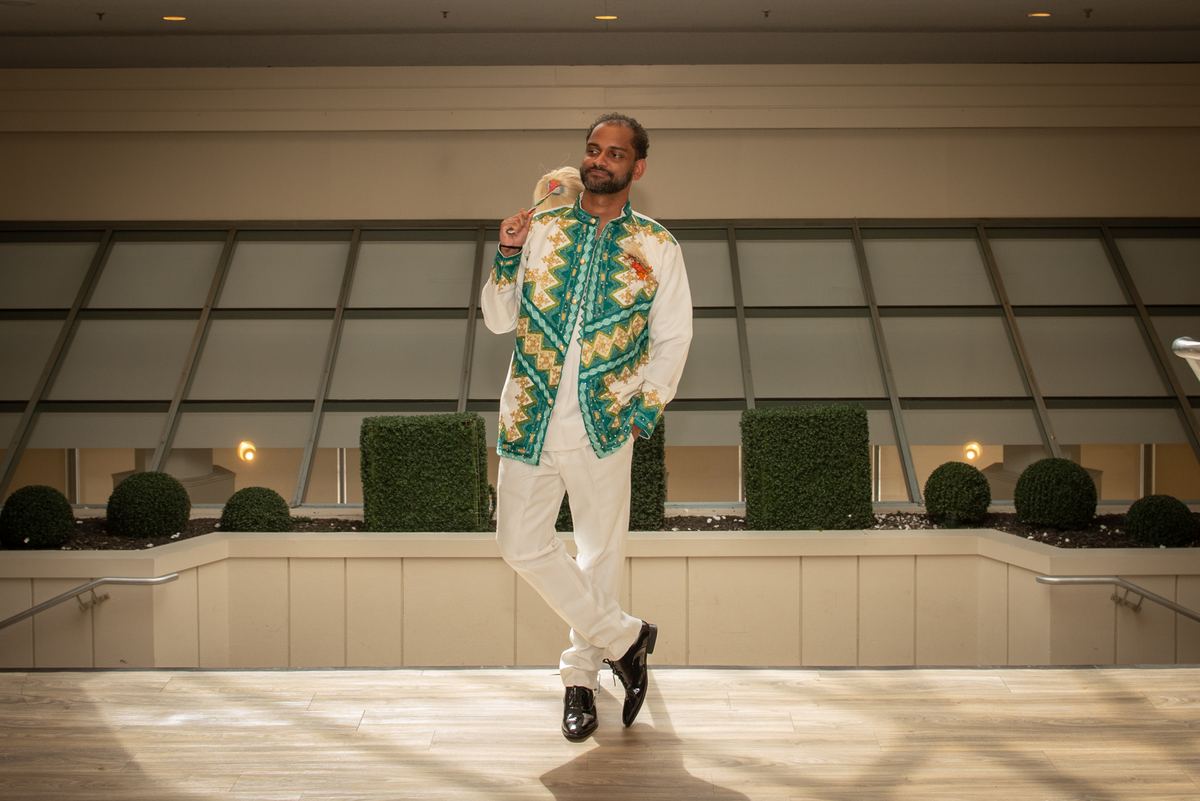 A man in a vibrant traditional jacket and white pants holds a ceremonial staff, standing confidently in sunlight.