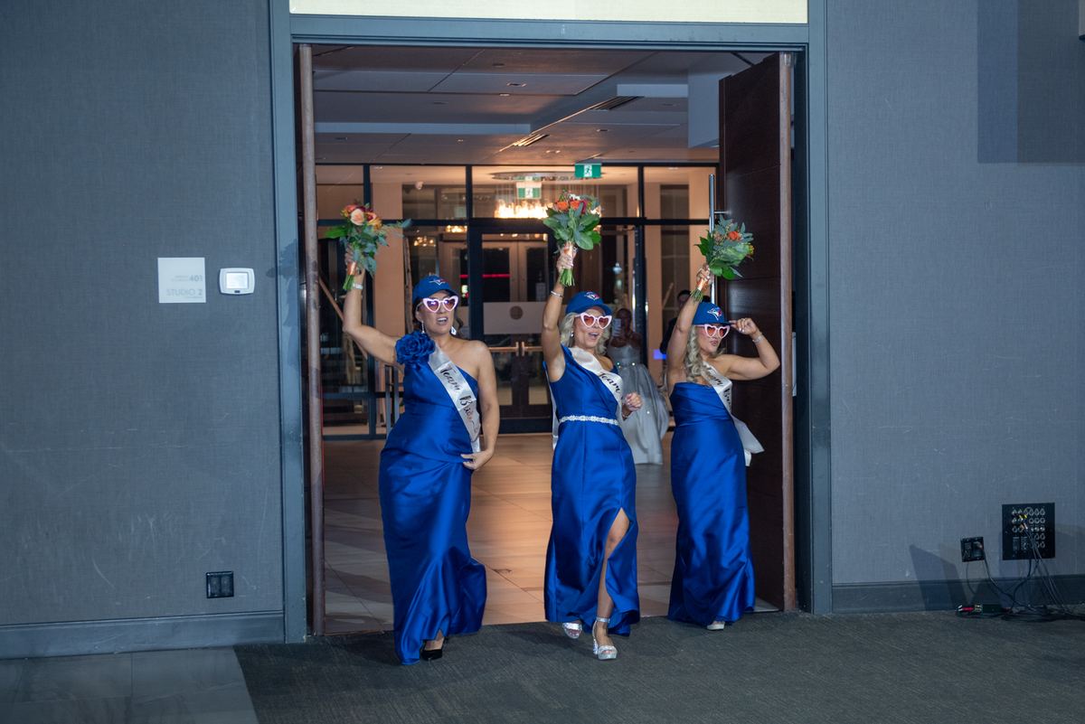 Three women in royal blue gowns and novelty sunglasses hold bouquets, exuding joy as they exit a doorway.
