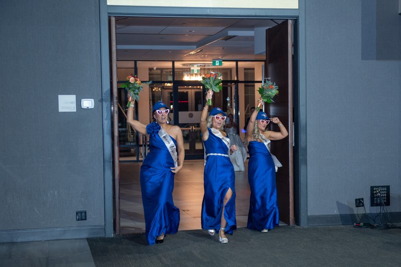 Three women in royal blue gowns and novelty sunglasses hold bouquets, exuding joy as they exit a doorway.