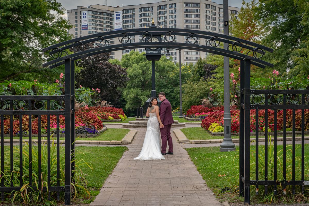 A bride and groom stand close under an ornate arch in a city park surrounded by flowers and skyscrapers.