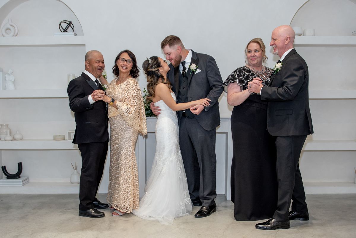 A bride and groom embrace warmly as their families gather in a circle around them, smiling and holding hands.