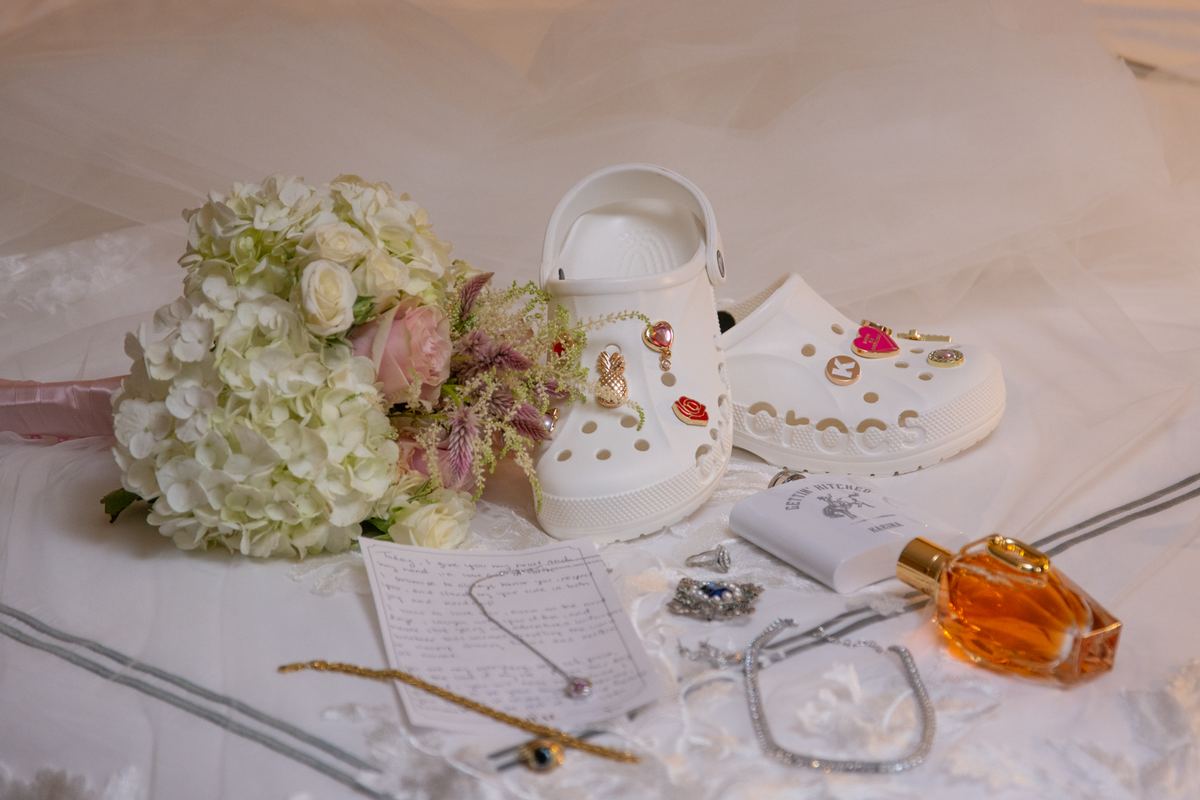 White Crocs with charms, flowers, jewelry, and perfume bottle arranged on tulle, creating a romantic bridal scene.