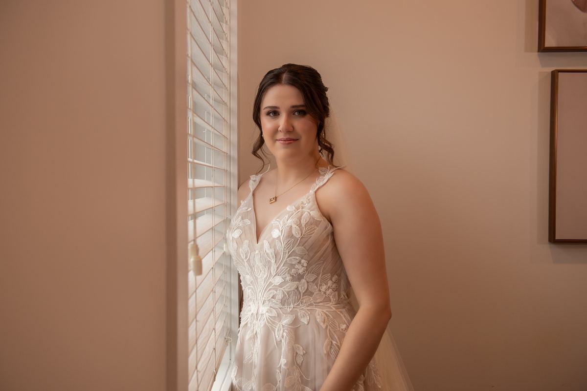 A bride stands calmly, her delicate lace gown illuminated by soft natural light filtering through vertical blinds.