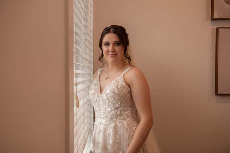 A bride stands calmly, her delicate lace gown illuminated by soft natural light filtering through vertical blinds.