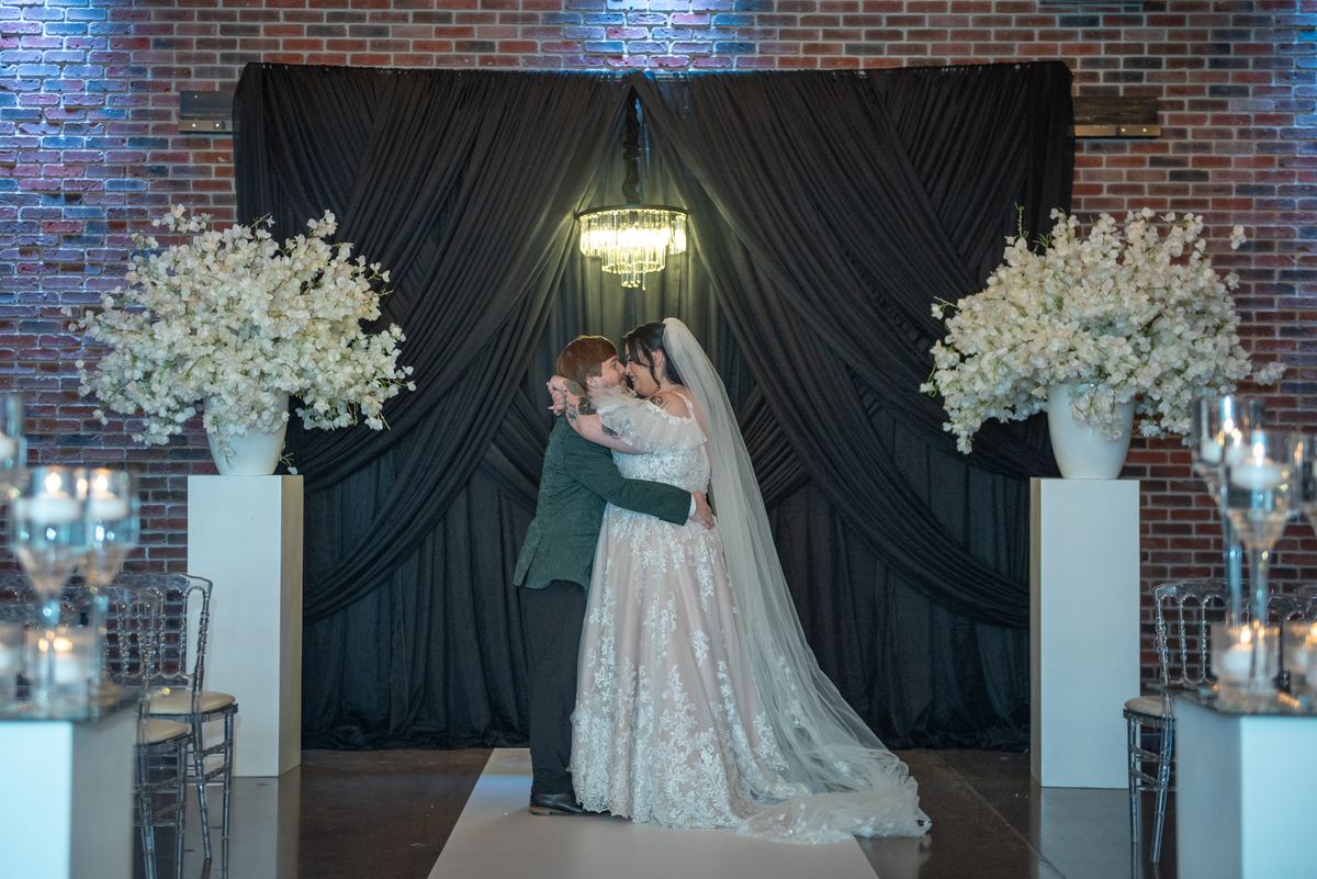 A bride and groom kiss at their wedding altar, surrounded by black drapery and white flowers, under warm chandelier light.