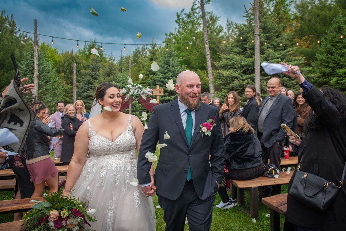 Bride and groom walk down the aisle surrounded by guests tossing petals, under string lights and trees.