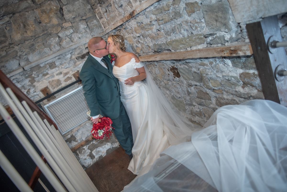 A bride and groom kiss in a rustic stone-walled room with their formal attire and the bride's veil visible.
