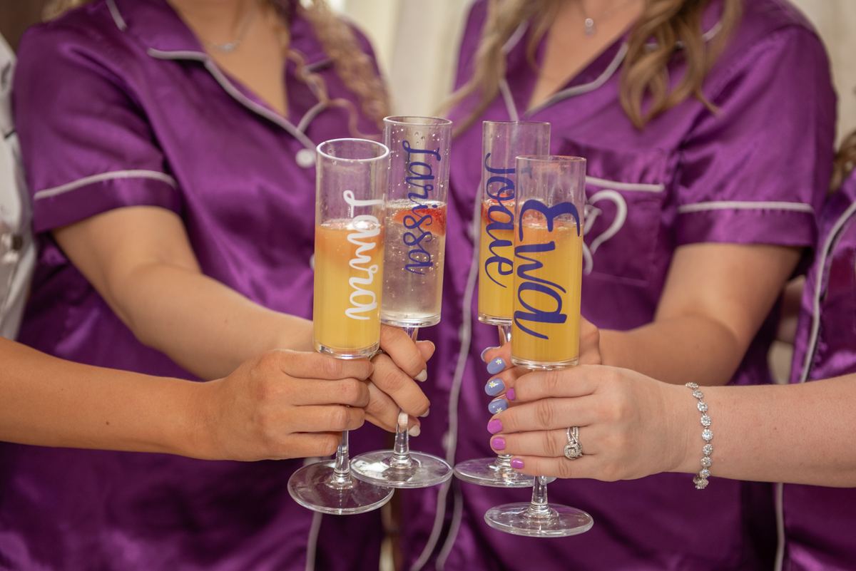 Four women in purple pajamas toast with champagne flutes etched with names, clinking glasses filled with bubbly drinks.