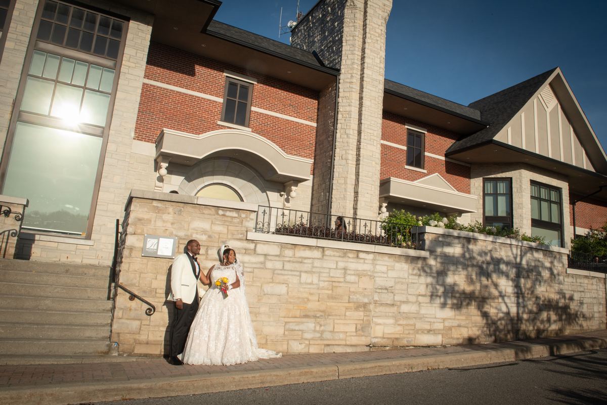 A newlywed couple in formal attire smiles beside a stone wall and brick building bathed in sunlight.