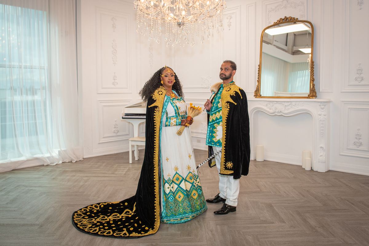 A couple in traditional Ethiopian attire stands in a white room illuminated by soft light from a chandelier.