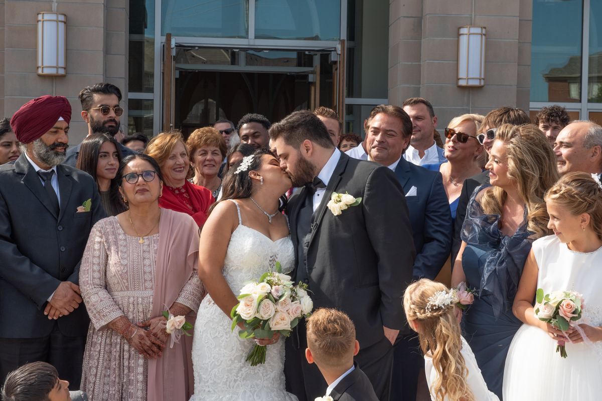A bride and groom kiss joyfully outside a modern building surrounded by smiling guests in formal attire.