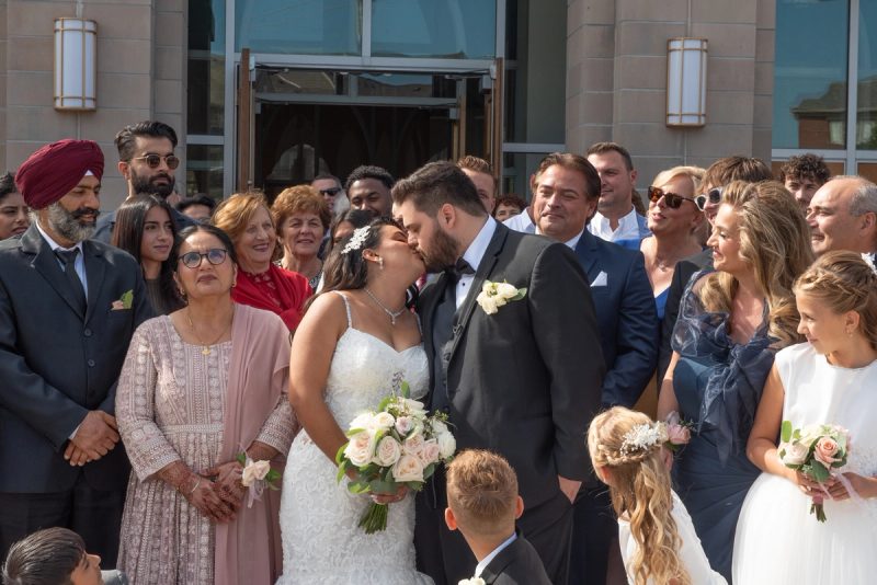 A bride and groom kiss joyfully outside a modern building surrounded by smiling guests in formal attire.